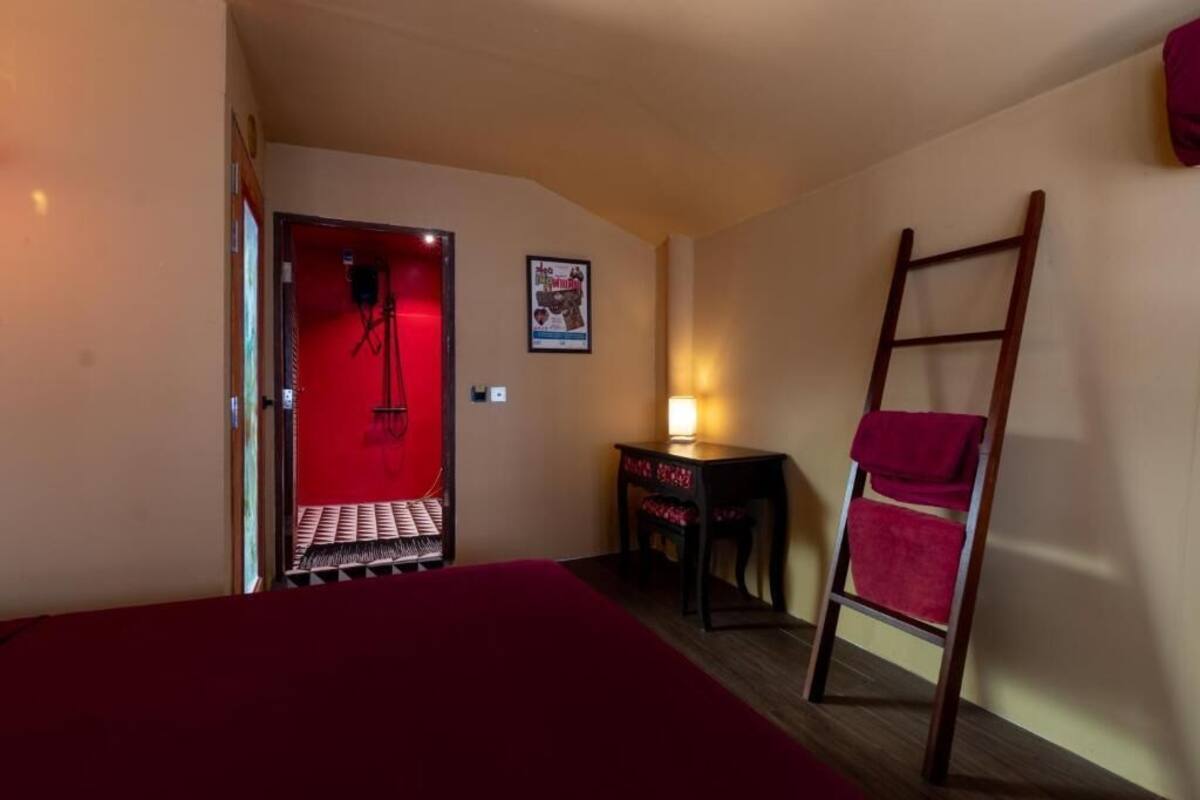 A bedroom with a red bedspread, a wooden ladder, a small table with a lamp, and a poster on the wall.