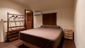 Ein Schlafzimmer mit einem Bett, einem Holzschrank, einer Kleiderstange, einem Fenster mit Vorhängen und einer kleinen Holzkiste.