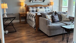 Un dormitorio con cama, un sofá gris, suelo de madera, ventilador de techo y ventana con cortinas.