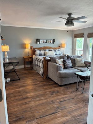 A bedroom with a bed, a gray sofa, a wooden floor, a ceiling fan, and a window with curtains. - Incredible lakefront spacious house with best view in town . Sleeps 15. (Granbury)