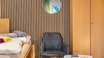 A room with a bed, a chair, a round mirror, and a small table with a book and glasses.