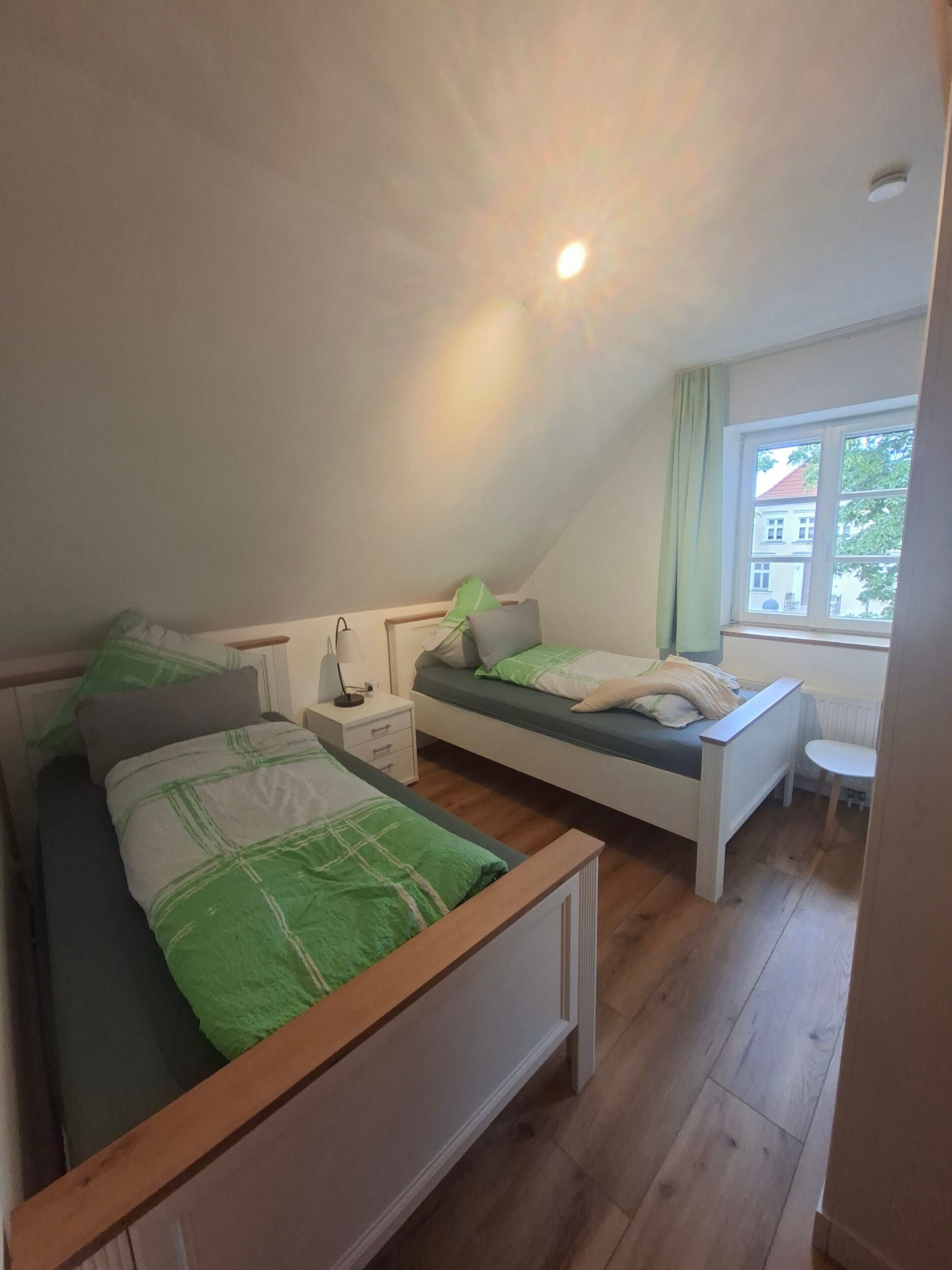 A bedroom with two beds, a nightstand, a lamp, and a window with a view of a building outside.. Zeitlos Wriezen 3 – Cozy Apartment for 4 with Garden, Parking, and Self-Check-in