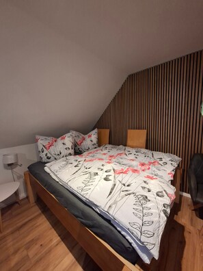A bedroom with a wooden bed, a bedside table, a chair, and a sloped ceiling with wooden paneling.