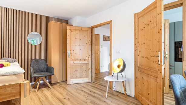 Una stanza con un letto, una sedia, uno specchio rotondo e porte in legno.