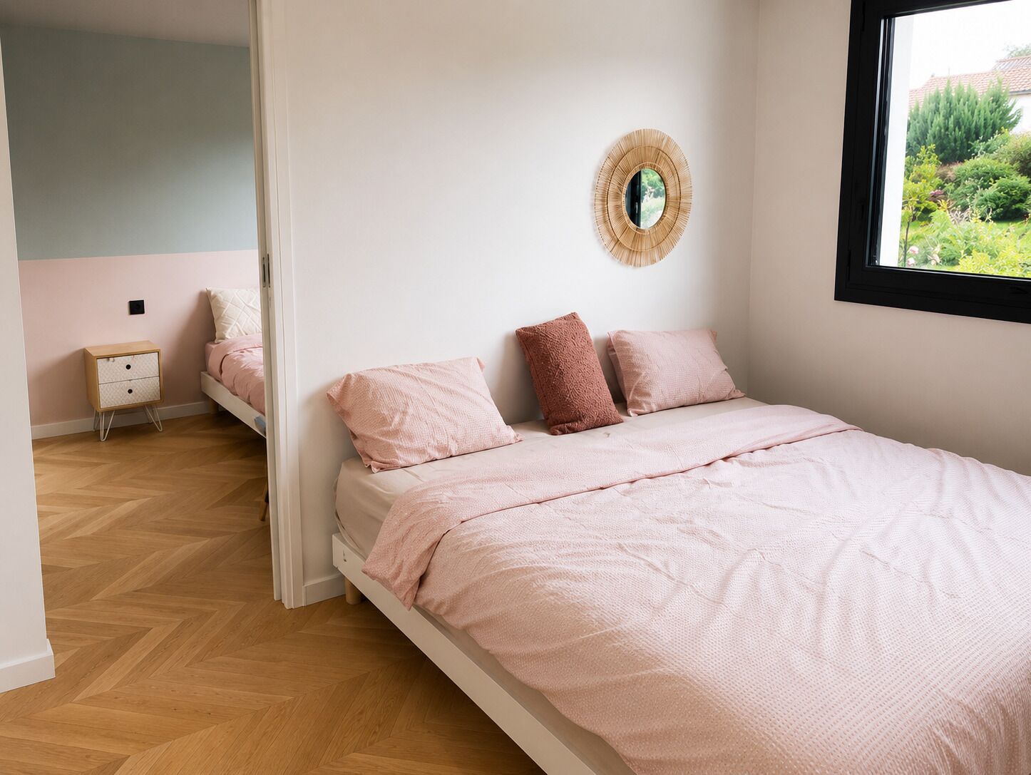 A bedroom with a bed, a bedside table, a mirror, and a window with a view of greenery.
