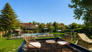 Eine Holzterrasse mit Korbstühlen und einem kleinen Tisch, mit Blick auf einen Swimmingpool und einen gepflegten Rasen.