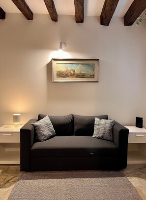 A modern living room with a dark sofa, two pillows, a framed painting, a side table with a lamp, and a speaker on a desk. - La Casetta—2-room apartment in the heart of Venice (Venezia)