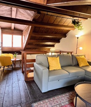 A cozy living room with a wooden ceiling, a grey sofa with yellow cushions, a yellow armchair, and a small table with a plant. - La Casetta—2-Bedroom Apartment in the Heart of Venice (Venezia)
