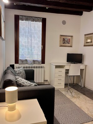 A modern living room with a sofa, a desk with a chair, a television, and a radiator. - La Casetta—2-Bedroom Apartment in the Heart of Venice (Venezia)