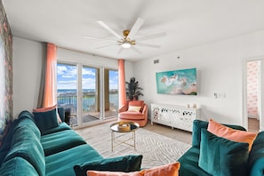 Ein Wohnzimmer mit türkisfarbenen Sofas, einem pinken Sessel, einem Deckenventilator, einem Fernseher und einem Balkon mit Blick.