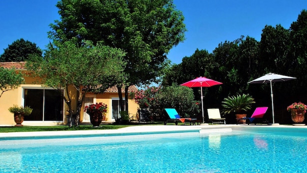 Piscina com espreguiçadeiras e guarda-sóis, cercada por árvores e um edifício.