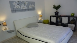 Quarto moderno com cama, luminárias de cabeceira, mesa de cabeceira, poltrona em formato de coração, planta em vaso e estante.