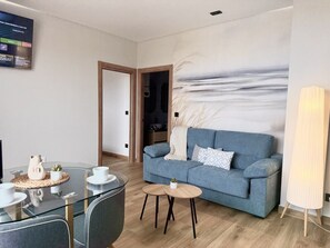 Un salon moderne avec un canapé bleu, une table à manger en verre agrémentée de tasses blanches et d’un pichet en céramique, une petite table ronde avec une plante, et un grand tableau abstrait accroché au mur.
