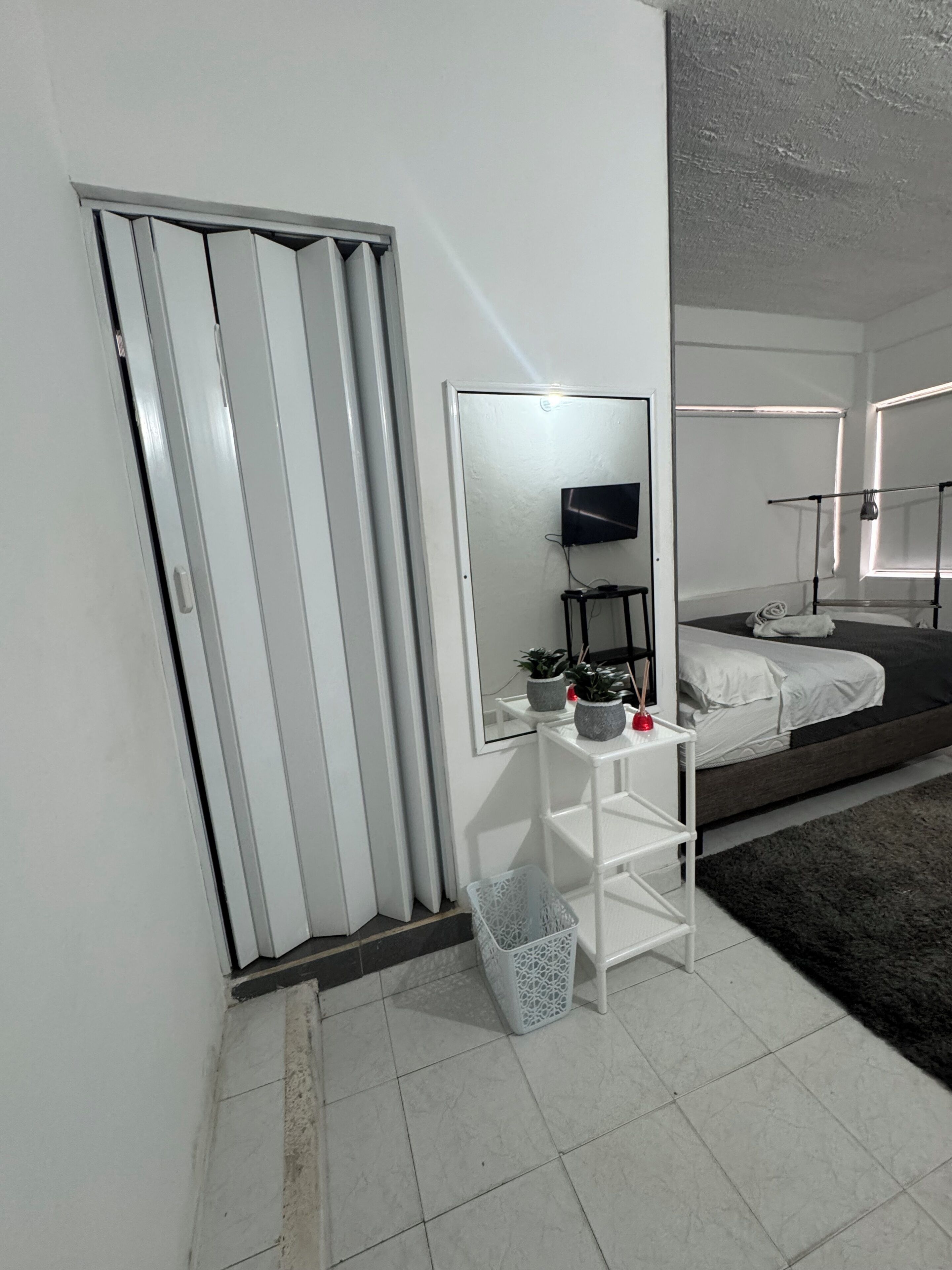 A modern bedroom with a bed, a bedside table with a mirror, a small plant, and a white wall unit.