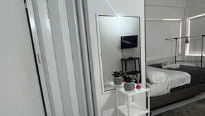 Un dormitorio moderno con una cama, una mesita de noche con espejo, una planta pequeña y un mueble blanco.