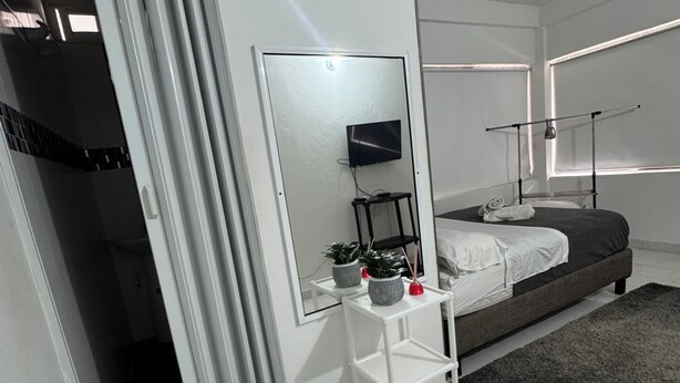Un dormitorio moderno con cama, mesita de noche con espejo, televisor y ventana con persianas.