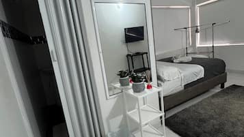 A modern bedroom with a bed, a bedside table with a mirror, a TV, and a window with blinds.