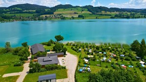 A lakeside property with a large body of water, a grassy area with tents, and a building with a dark roof. - Cubes am Irrsee - Self Check-In (Fischhof)