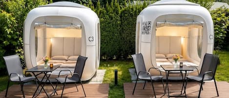 Two outdoor sleeping pods labeled C1 and C3, each with a table and chairs, set on a wooden deck with grass in the background.