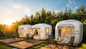 Three modern, white, dome-shaped tents labeled C1, C2, and C3, each with a small table and chairs, set on a wooden deck in a grassy area with a dense hedge in the background. - Cubes am Irrsee - Self Check-In (Fischhof)