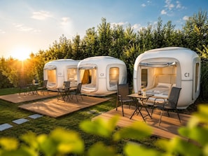Three modern, white, dome-shaped tents labeled C1, C2, and C3, each with a small table and chairs, set on a wooden deck in a grassy area with a dense hedge in the background. - Cubes am Irrsee - Self Check-In (Fischhof)