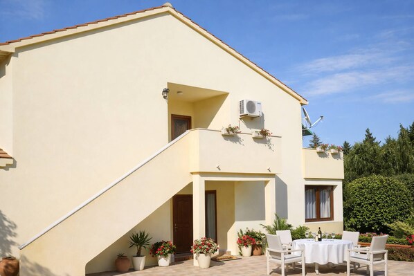 Ein zweistöckiges Gebäude mit flachem Dach, einem Balkon mit Topfpflanzen, einer Treppe und einer Terrasse mit Tisch und Stühlen.