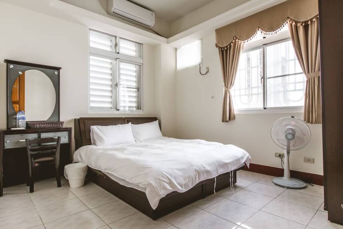 Un dormitorio con cama, escritorio con silla, espejo, ventana con cortinas y ventilador de pie.
