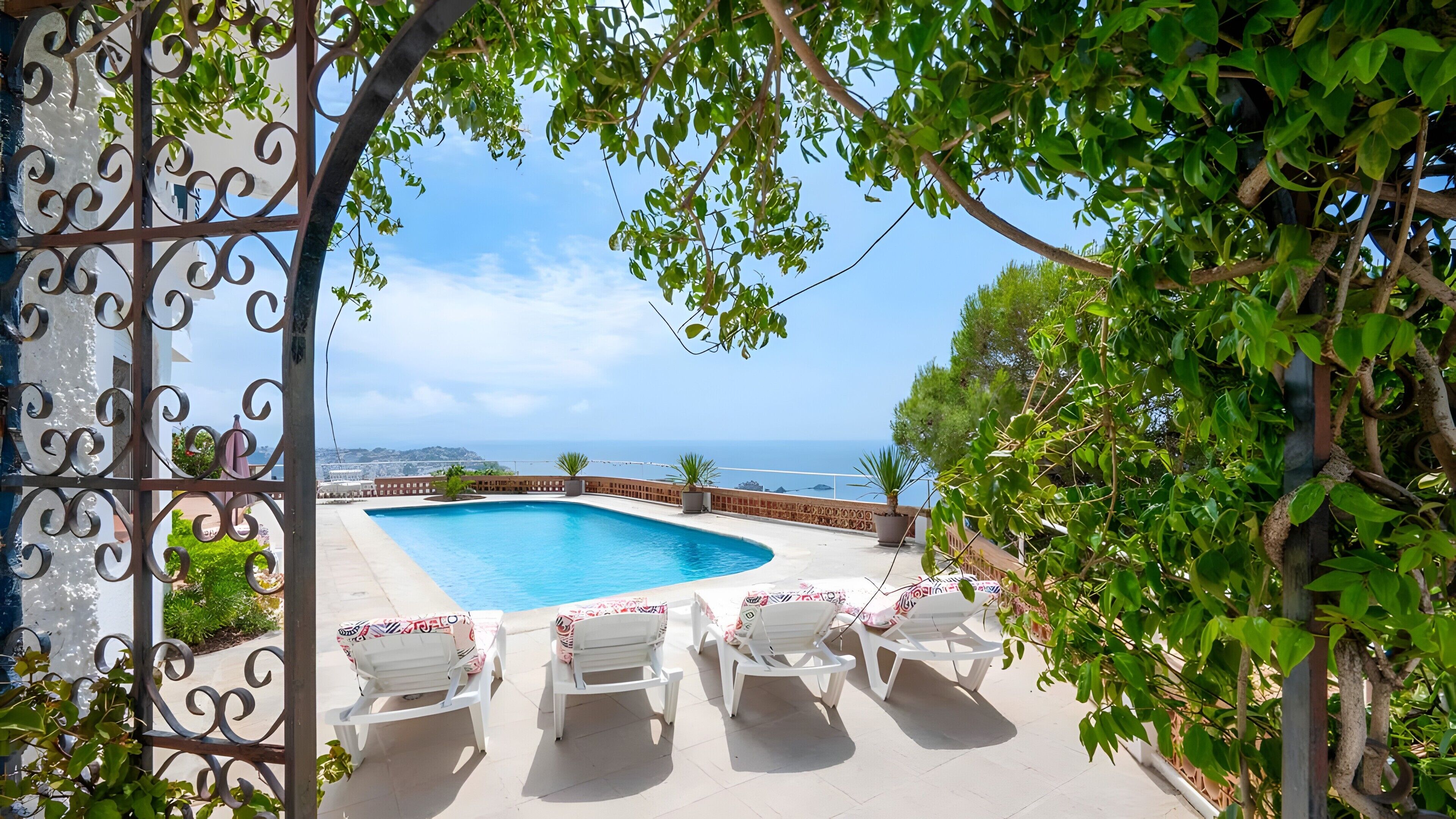 A swimming pool with lounge chairs, surrounded by greenery and overlooking a coastal view.