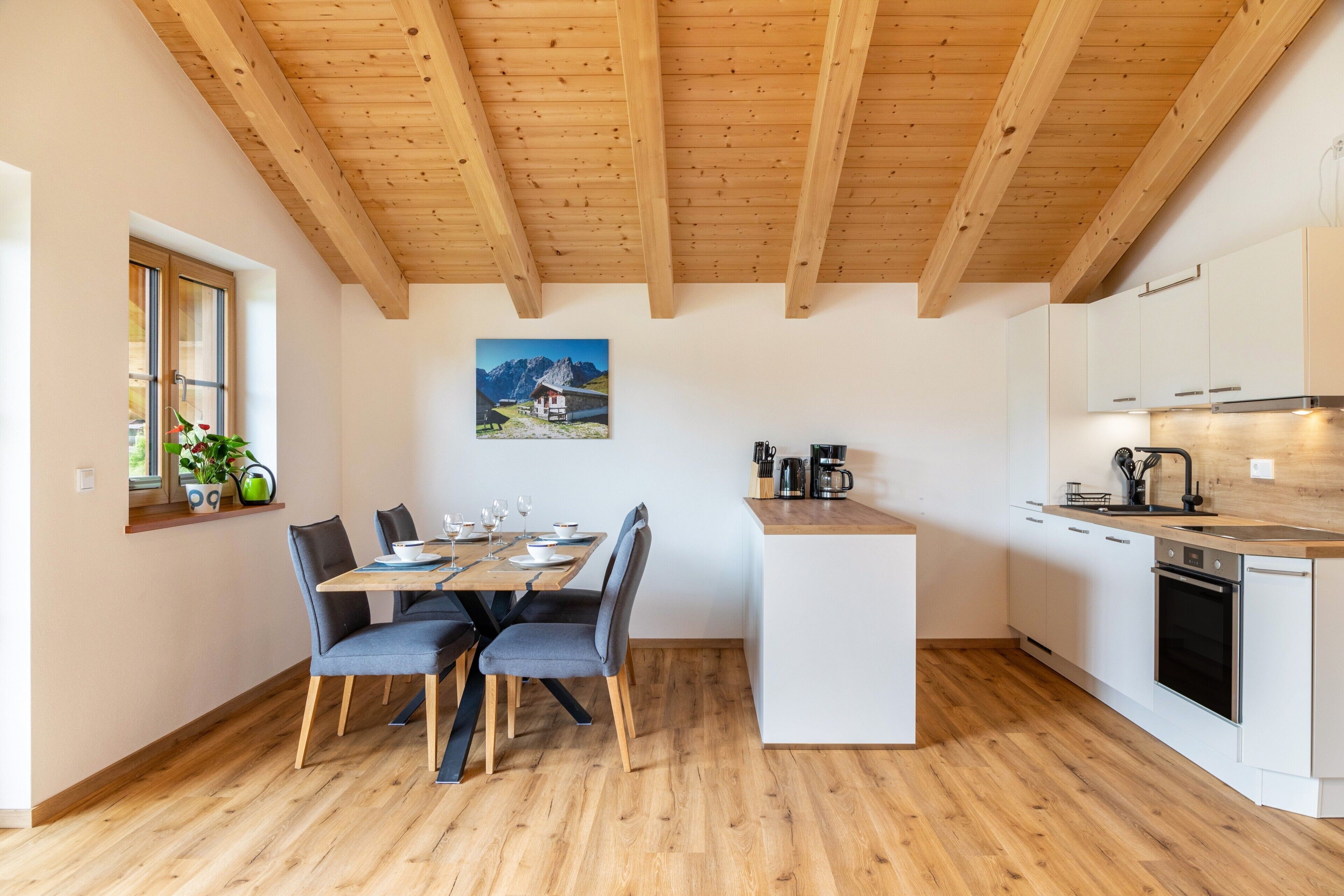 Una cocina moderna con vigas de madera, un comedor con mesa y sillas, y un ventanal con vistas a montañas.