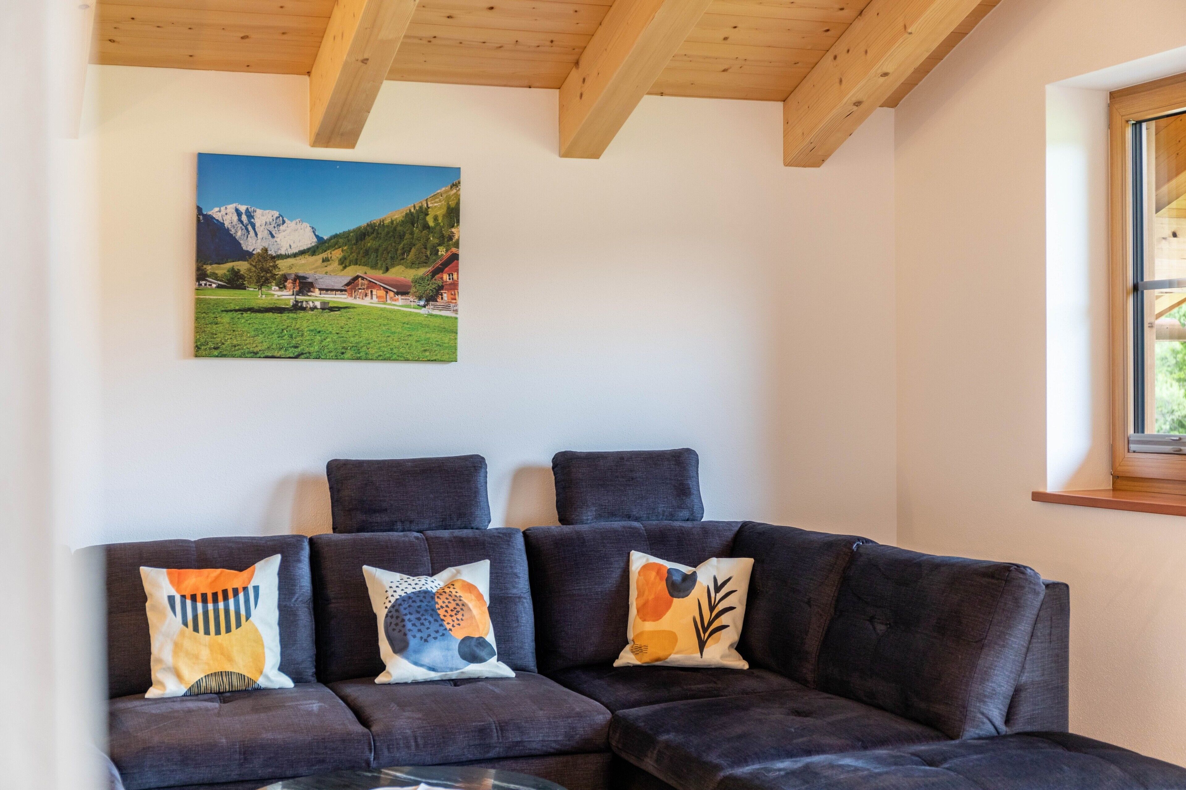 A sectional sofa with three cushions, a wooden ceiling, a framed landscape painting, and a window with a view of greenery.