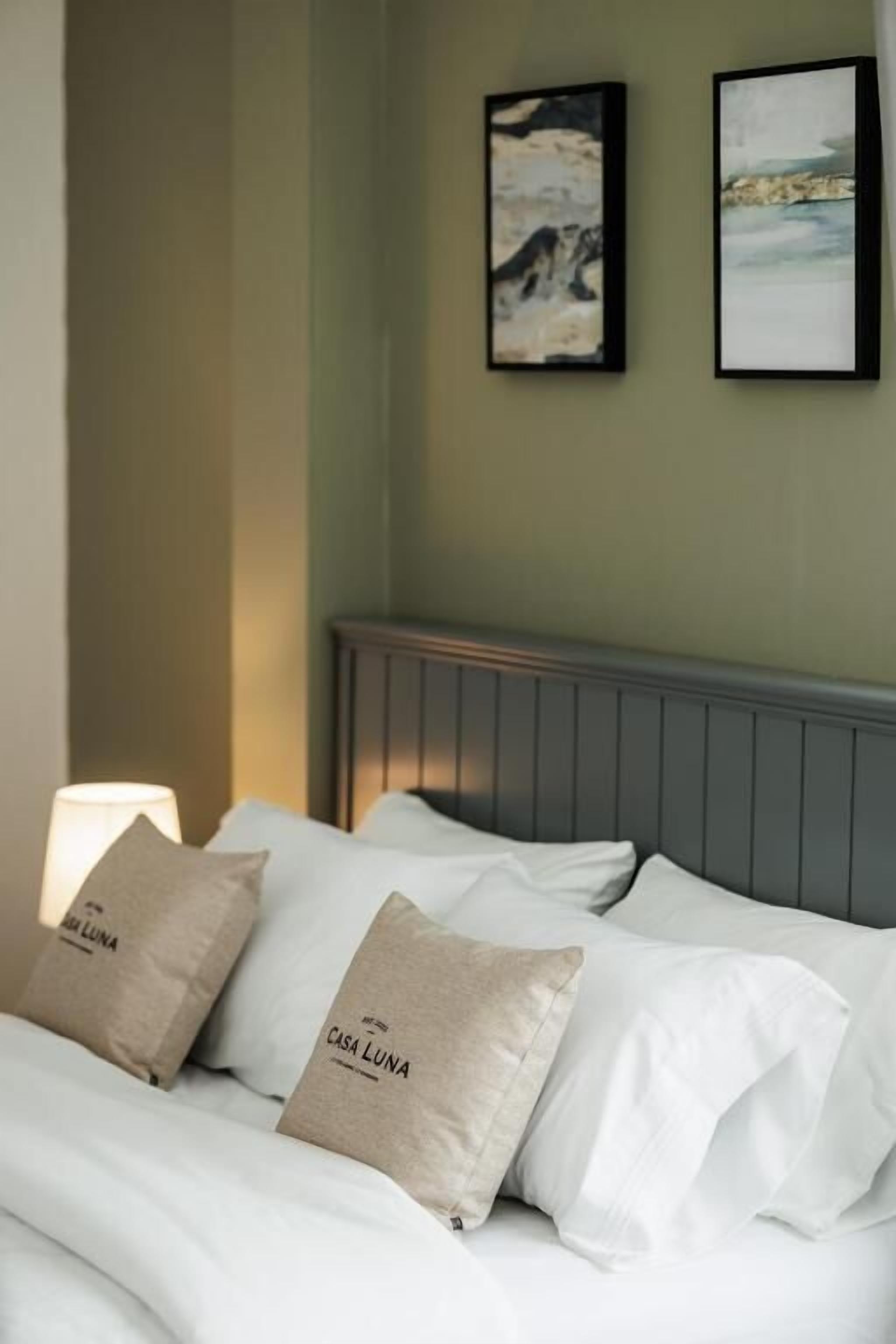 A bed with white bedding and two pillows, a bedside lamp, and two framed pictures on the wall.