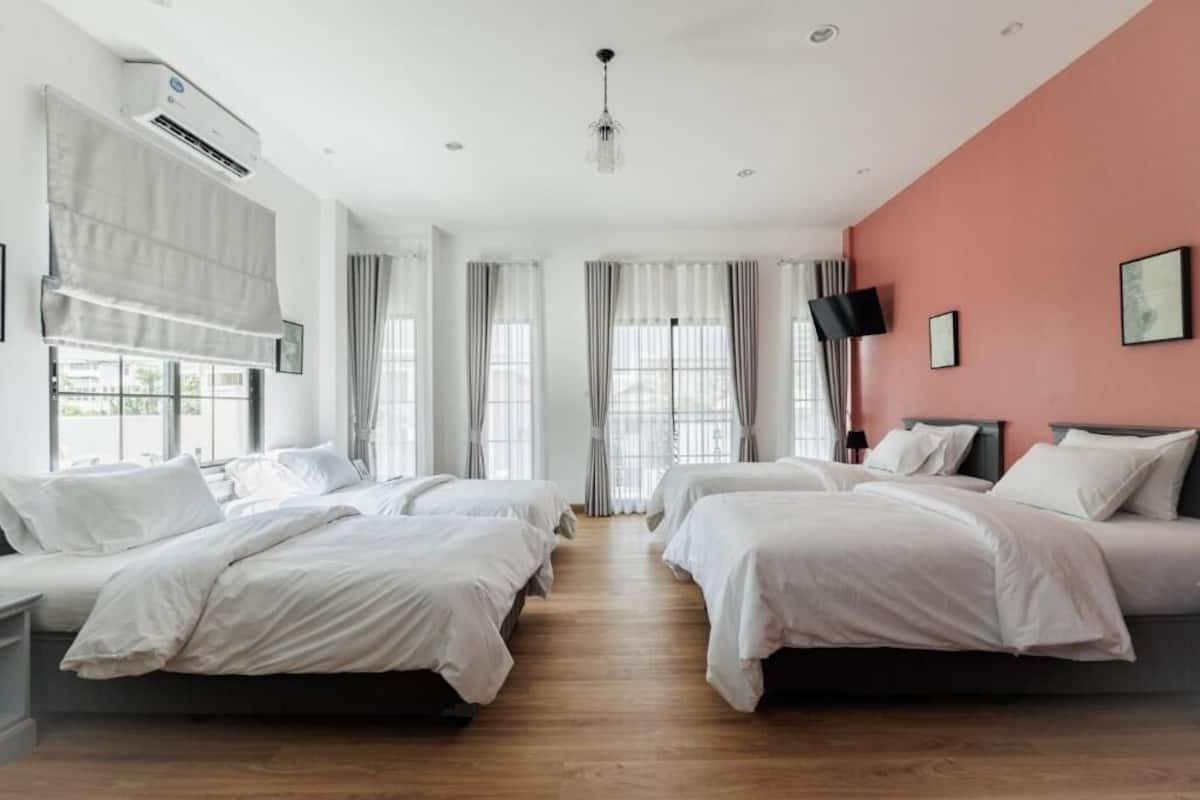 A hotel room with four beds, large windows with curtains, a flat-screen TV, and an air conditioning unit.