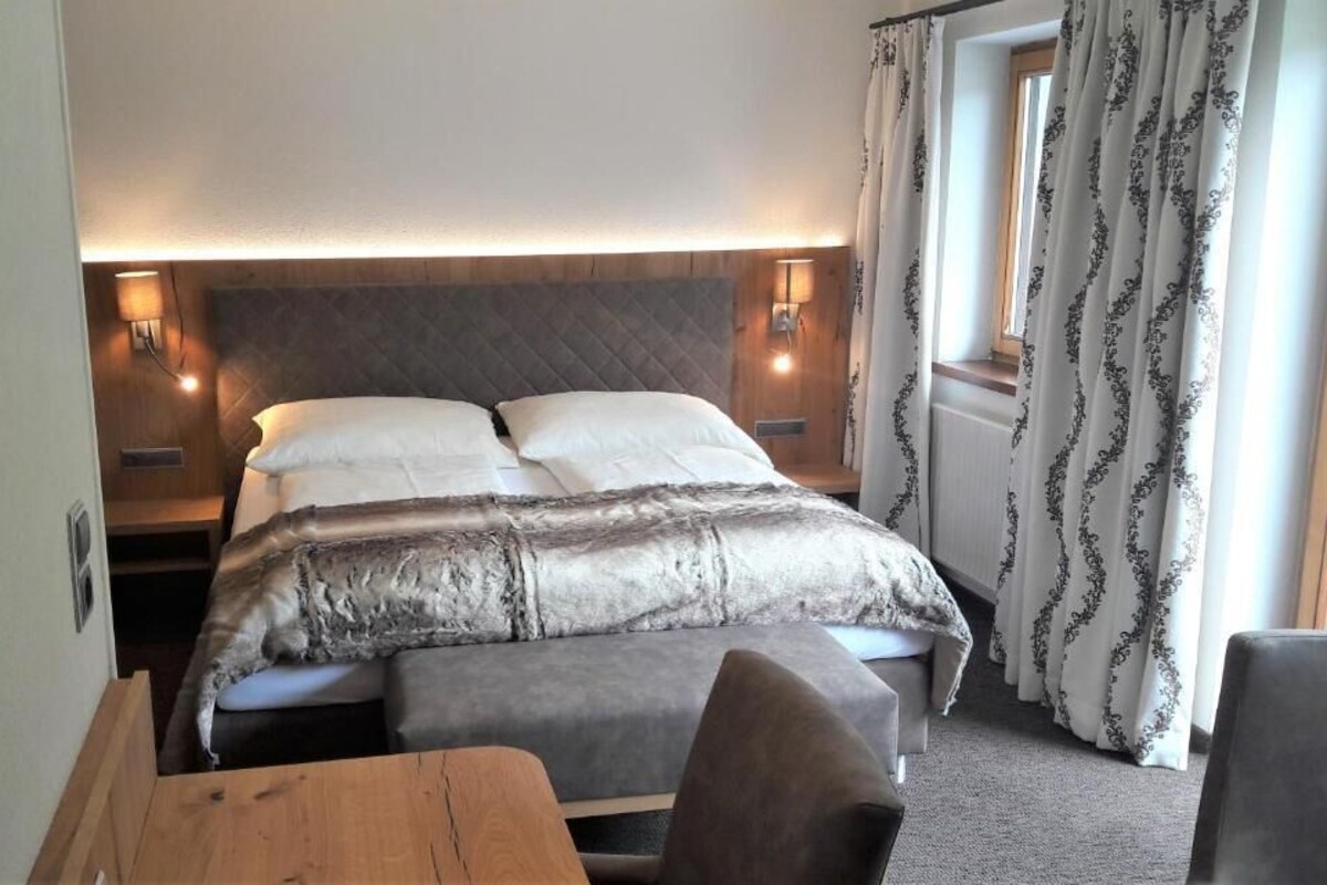 A bedroom with a bed, bedside lamps, a wooden nightstand, a bench, and a window with curtains.