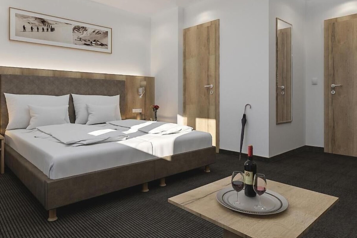 A hotel room with a bed, a bedside table, a framed picture on the wall, and a table with a bottle and glasses.