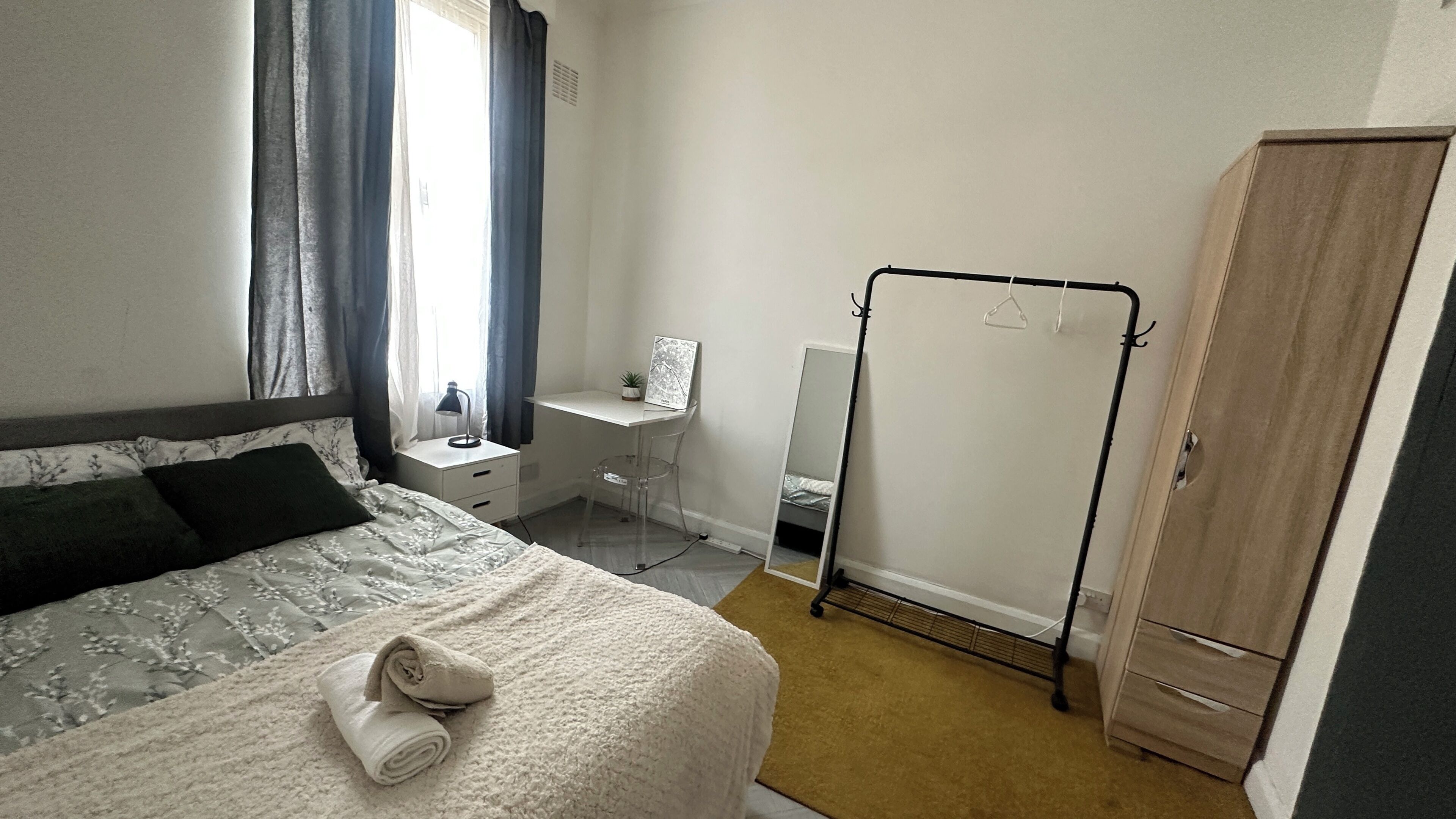 A bedroom with a bed, a desk, a mirror, a wardrobe, and a window with curtains.