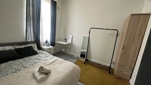 A bedroom with a bed, a desk, a mirror, a wardrobe, and a window with curtains.