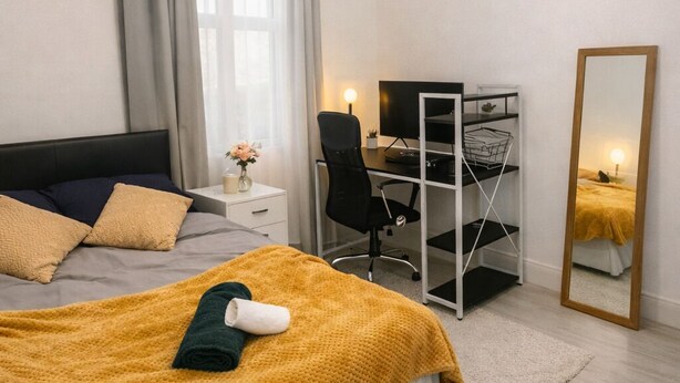 A bedroom with a bed, a desk, a chair, a mirror, and a window with curtains.