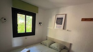 A single bed with pillows and folded towels, a window with a view of greenery, a wall-mounted shelf with a framed item, and a thermostat on the wall. - Charming 2-room apartment with magnificent sea view (Grosseto-Prugna)