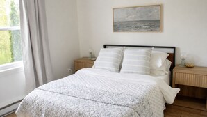 Ein Schlafzimmer mit einem Bett, zwei Nachttischen, einem Schminktisch, einem Gemälde und einem Fenster mit Vorhängen.