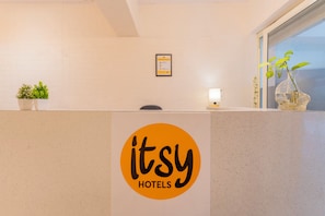 A reception desk with a logo, a potted plant, a framed certificate, and a window with a view. - Itsy Hotels Cozy Retreat - Near Mysore Palace & Zoo (Mysore)