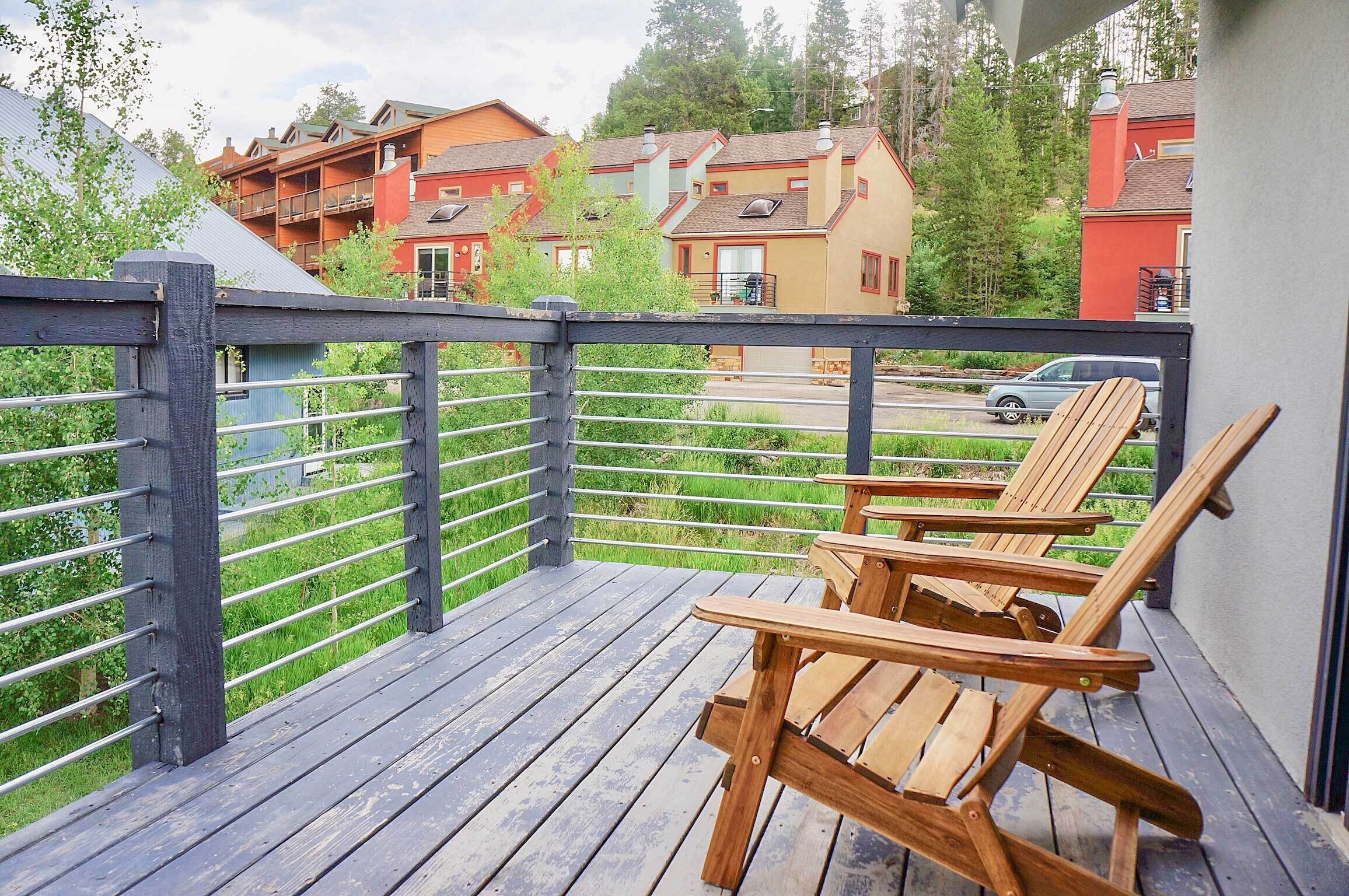 Una terraza de madera con tres sillas de madera, barandilla de metal y vista a edificios residenciales al fondo.