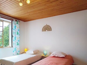 Ein Zimmer mit zwei Betten, Holzboden, einem Fenster mit Vorhängen, einem Wandventilator und einem kleinen Tisch mit Lampe.