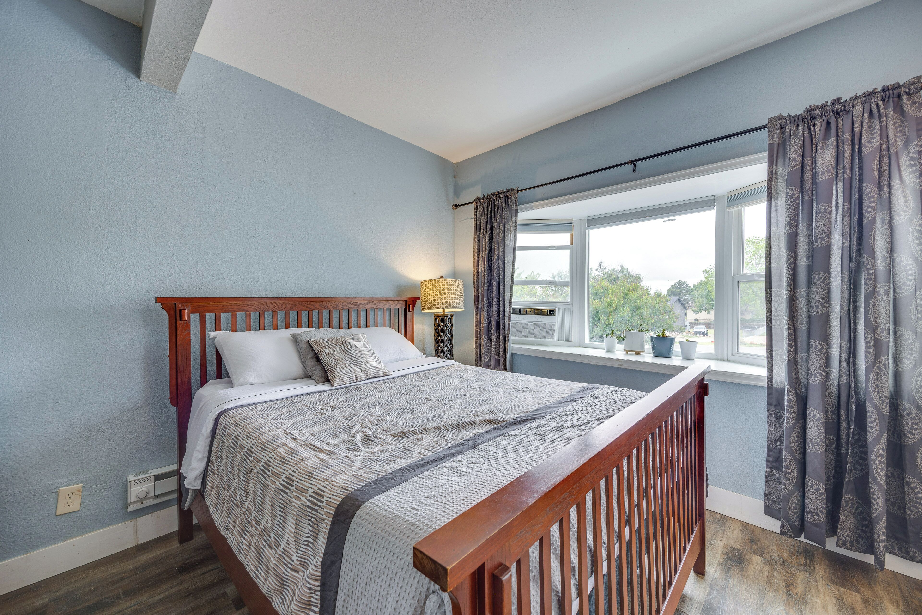Un dormitorio con cama de madera, ventana con cortinas, lámpara de noche y vistas a zonas verdes.