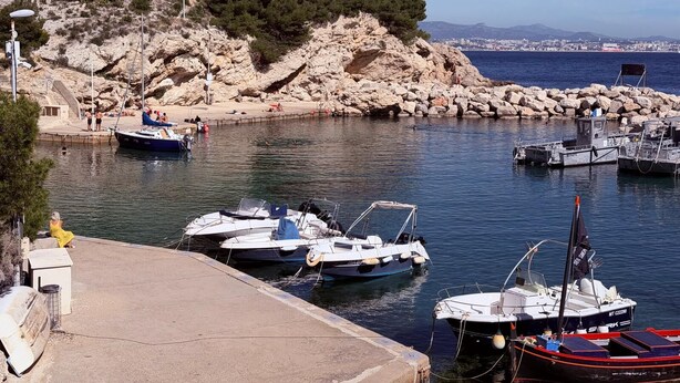 Un port de plaisance situé sur la côte, avec des bateaux amarrés, un terrain rocheux et une vue dégagée sur la ville au loin.