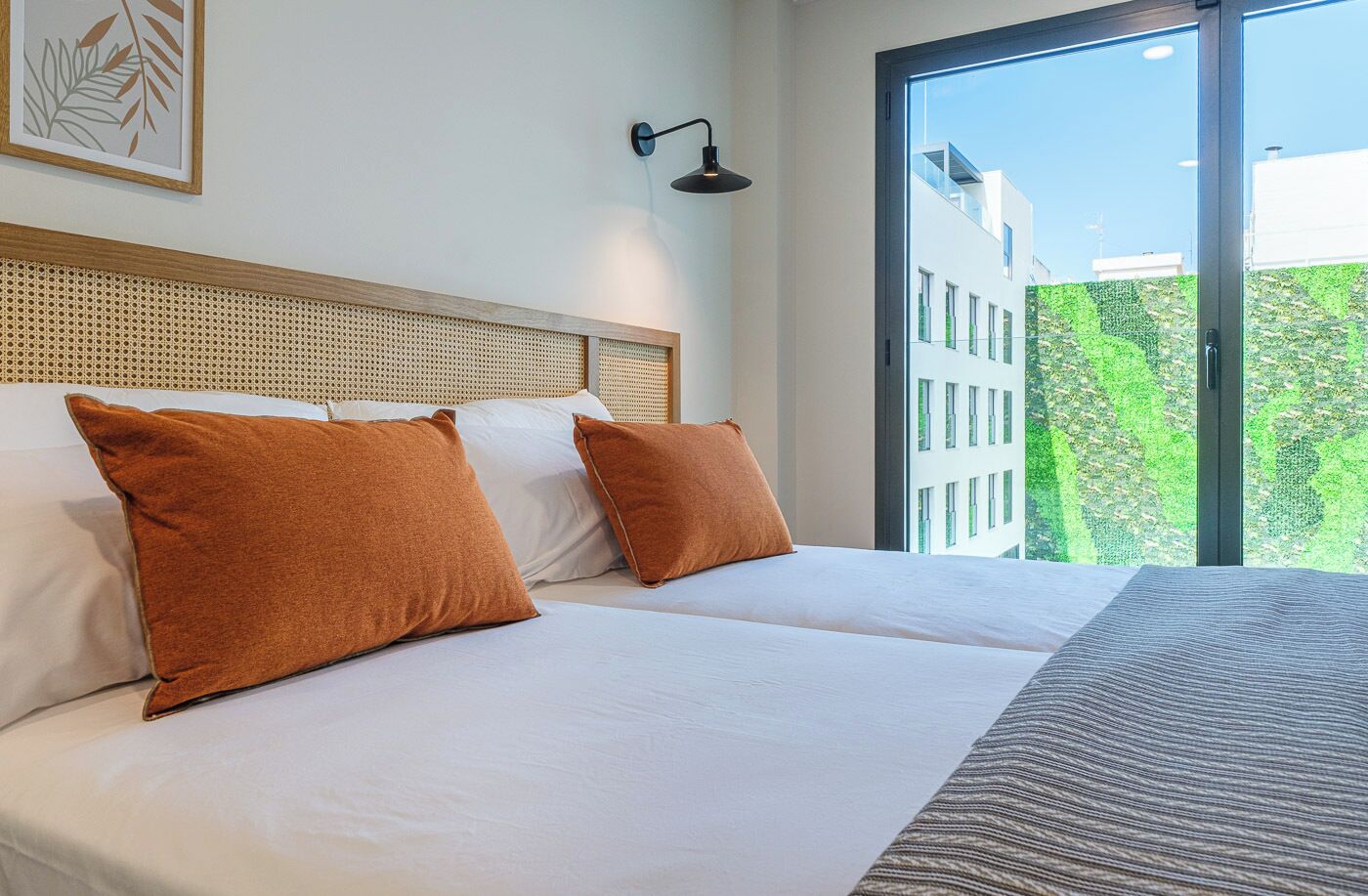 Habitación de hotel con una cama grande, dos almohadas, cabecero, luz de pared y ventana con vistas a edificios y vegetación.