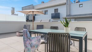 Una terraza en la azotea con una mesa de vidrio, sillas, una maceta y un paraguas grande.