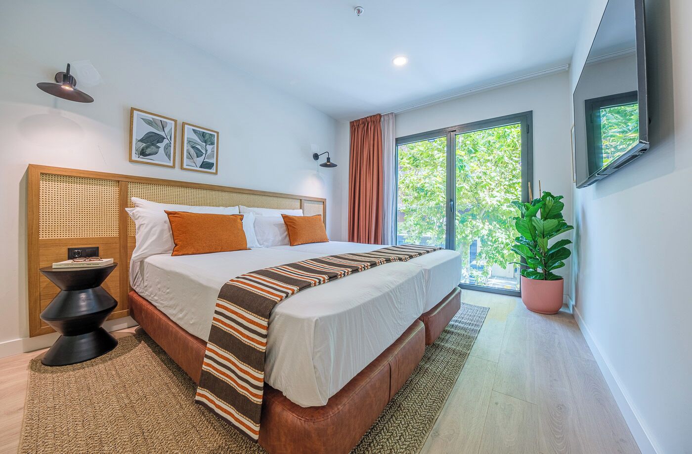 Une chambre moderne avec un grand lit, une tête de lit en bois, une table de chevet, une plante en pot et une grande fenêtre.