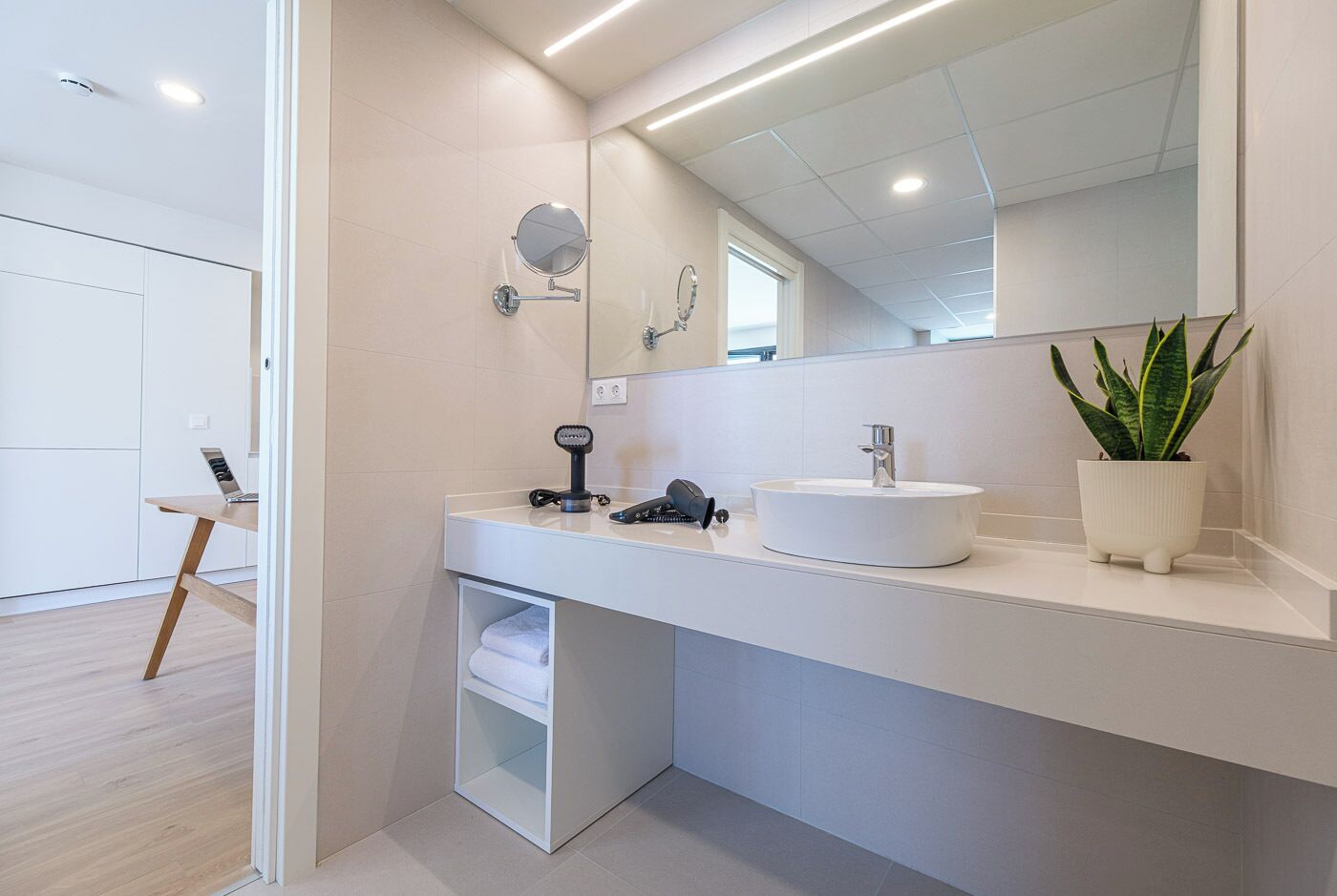 Un baño moderno con tocador blanco, espejo redondo, lavamanos y planta en maceta.