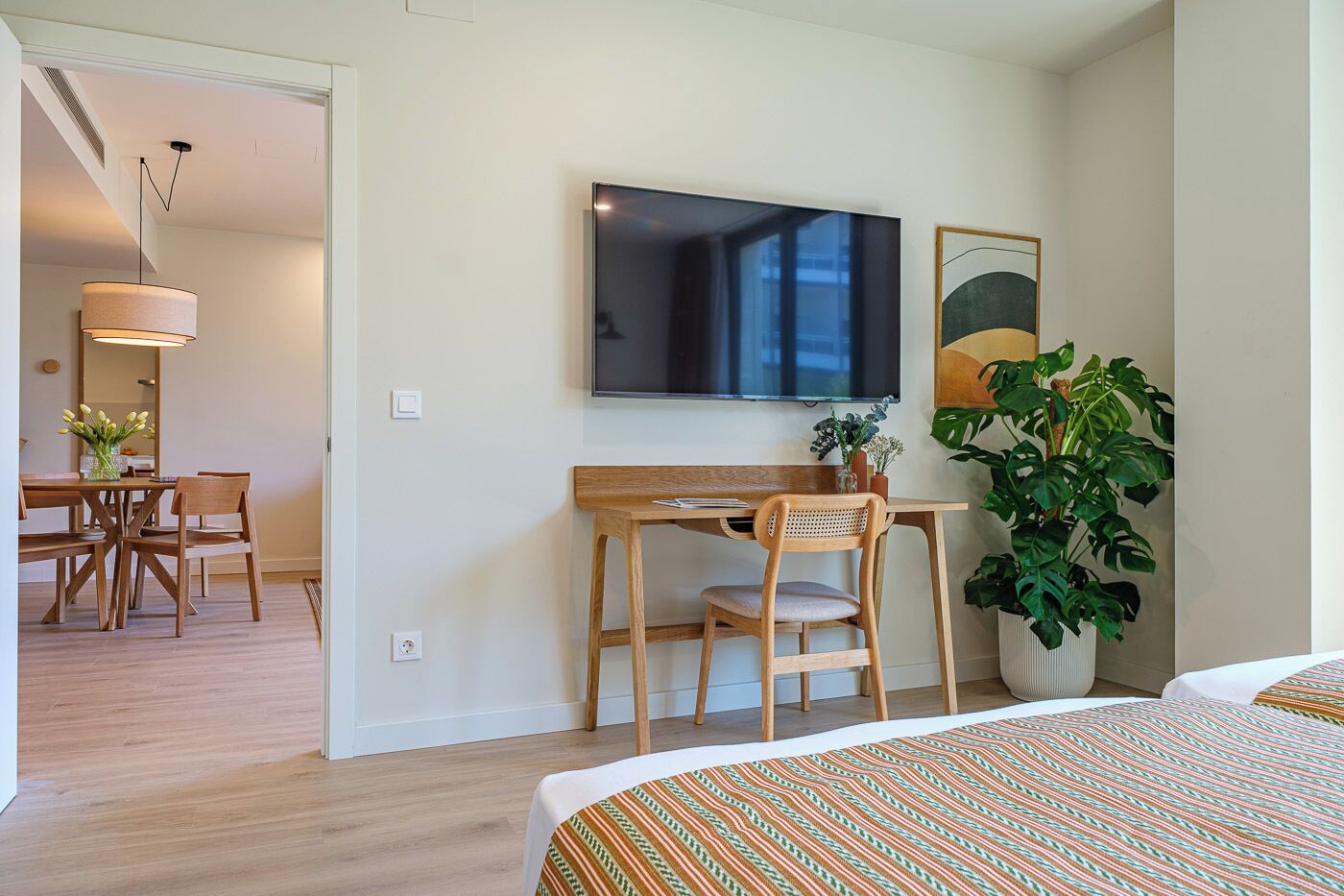 Habitación de hotel con cama, escritorio y silla, televisor, planta y área de comedor con mesa y sillas.