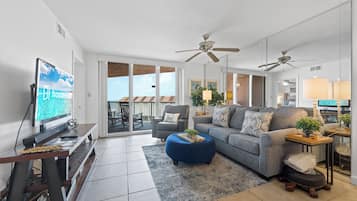 A modern living room with a sofa, ottoman, TV, ceiling fans, and a balcony with a view.